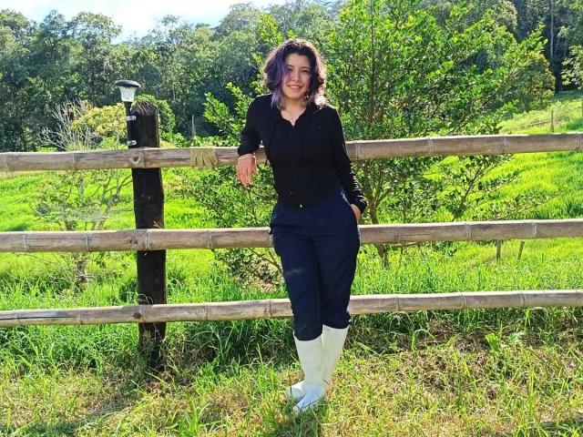 DarckVelvet with wavy brown hair, wearing a black shirt, navy pants, and white boots, leans on a wooden fence in a sunny, green grassy field.