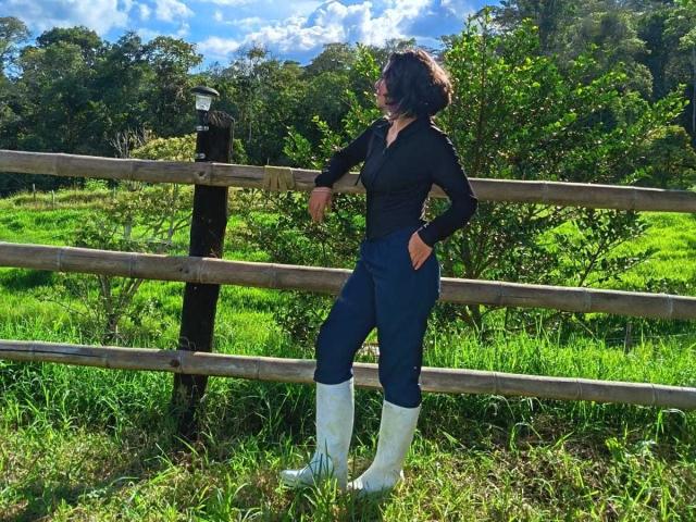 DarckVelvet stands in a grassy field, leaning on a wooden fence. She's wearing a black top, blue jeans, and white knee boots. She's looking to the side with a smile.