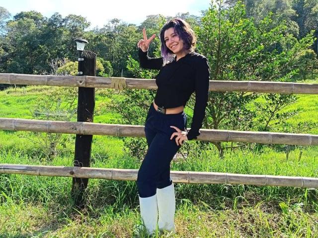 DarckVelvet stands in front of a wooden fence, wearing a black top, navy pants, and white boots, flashing a peace sign, with curly brown hair and a smiling face.