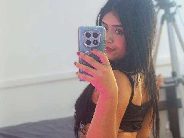DiamondSweet with long black hair, nose piercing, wearing a black sports bra, taking a selfie with a blue phone, showing her small tits, in a softly lit room.