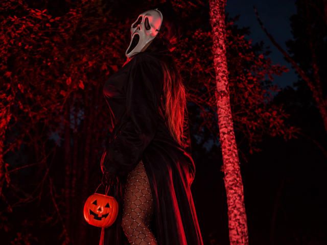 EmmaEbony with long red hair, black mask, black dress, fishnet stockings, holding glowing pumpkin bucket, standing in dark forest, red lighting, showing her ass.