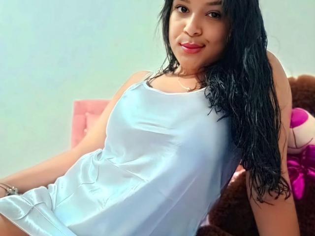 MiaBlakeX with long black wet hair, wearing a shiny light blue dress, sitting on a pink chair, smiling, showing her small tits, with a brown stuffed toy behind.