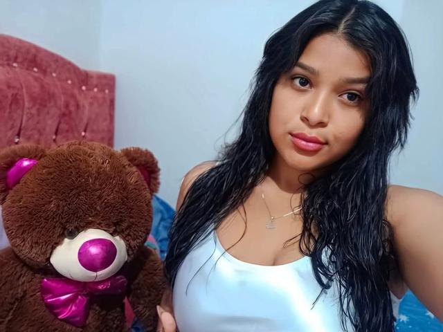 MiaBlakeX with wet black hair, wearing a white tank top, holding a brown teddy bear with a pink nose and bow, looking at the camera with pink lips.
