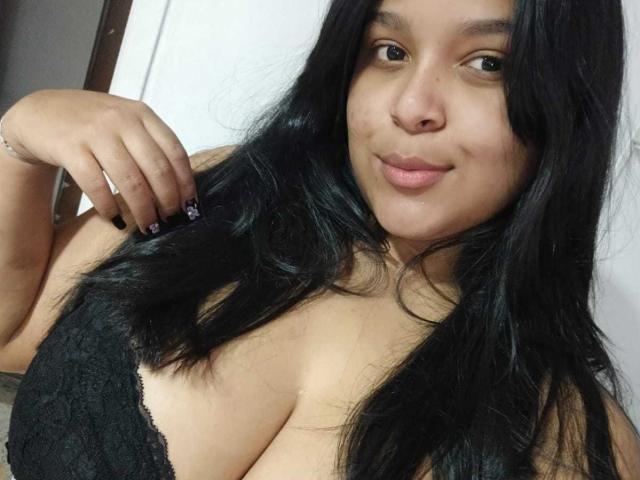 MiaBlakeX with long black hair, brown eyes, and a smile, wearing a black lace top, showing her big tits, holding her hair with black nails, and a silver bracelet.