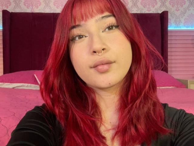 HaileyParck with red hair, nose ring, and black shirt, smiling at the camera, pink bed in background, light skin, subtle makeup, and relaxed expression.
