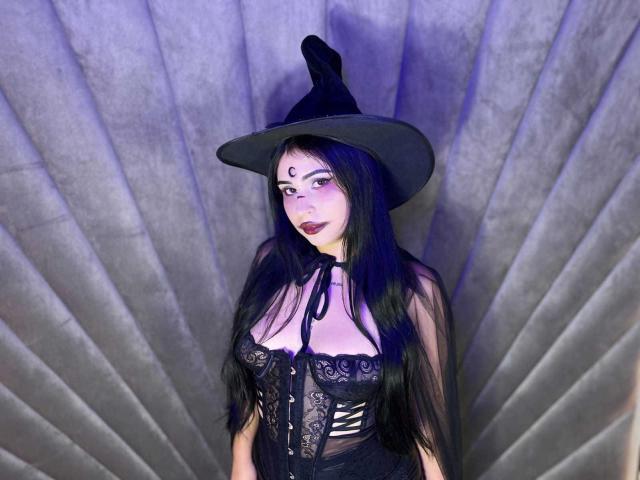HannahBecker with long black hair, wearing a witch hat and black lace corset, showing her tits, pale skin, purple lipstick, and a forehead gem, against a fan-like backdrop.
