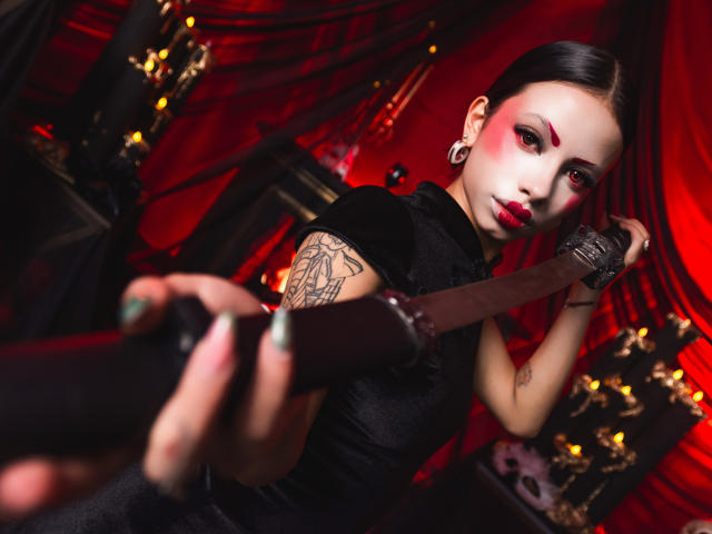 Kazumii with black hair, red makeup, and earrings, holding a knife, wearing a black top, showing a tattoo on her arm, and looking at the camera. Her tits are small.