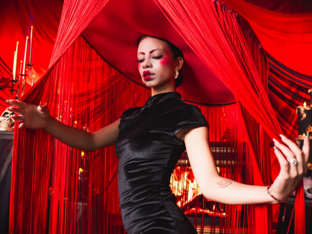 Kazumii in a tight black velvet dress, red face paint, and red hat, holding skull, with red curtains, her ass slightly raised, looking sexy, eyes closed, lips parted.
