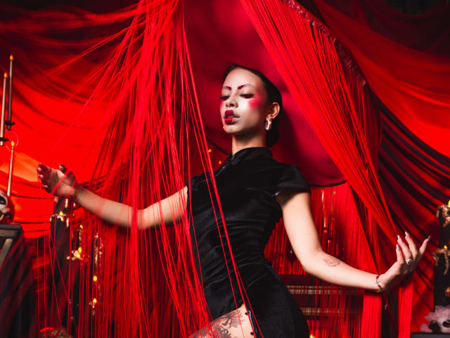 Kazumii with red face paint, black velvet dress, and tattoo on thigh, standing in red curtain, holding strings, arms out, looking seductive, red lipstick, confident, sexy.
