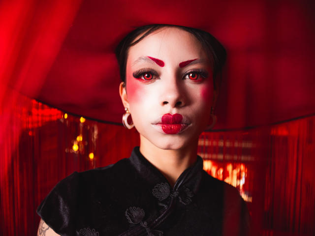 Kazumii has red makeup, bold eyebrows, and red lipstick. She's wearing a black cheongram with large hoop earrings. Her skin is pale, and she's looking straight at the camera.