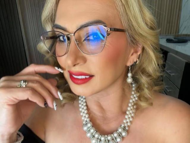 MisseScarleth with blonde curls, red lipstick, pearl necklace, and glasses, touching her cheek, showing off her white nails and pearl earrings, with a blue reflection in her glasses.