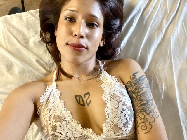 KiaraHernandezX lying on a bed, wearing a white lace bralette, showing small tits, tattoo above her chest, tattoo on her right arm, brown hair, subtle smile.
