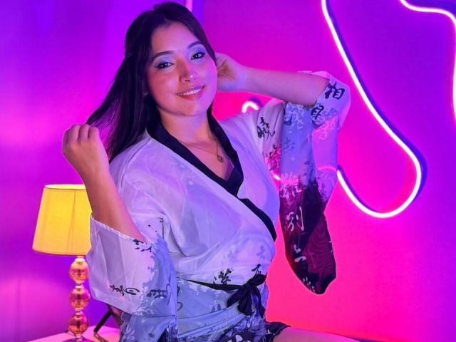  AlexandraZanis with long dark hair, wearing a light blue floral kimono, black trim, and a black belt, smiling, holding her hair, next to a yellow lamp.