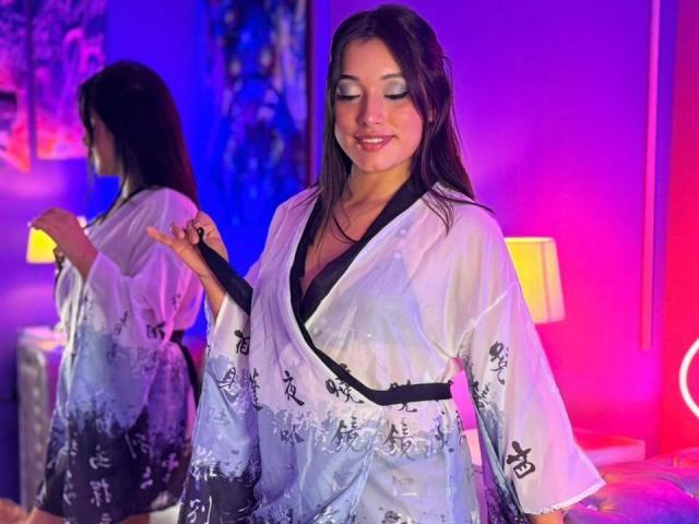 Alexandra Zanis, con cabello largo y oscuro, vestida con un kimono blanco translúcido con estampados florales negros, sonriendo, mostrando sus pechos, de pie en una habitación con iluminación neón púrpura y rosa..