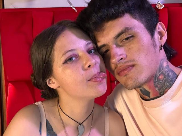 GraceAndTony and Tony with their tongues out, GraceAndTony with small tits, wearing a gray tank top, Tony with tattoos, black hair, and ear piercings, on red booth.