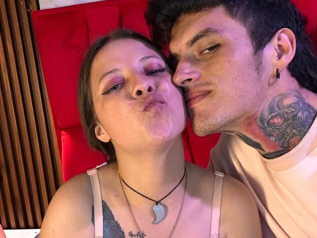 GraceAndTony with puffy lips, next to Tony with tattoos, wearing a pink shirt, kissing her cheek. She's in a grey top, he's earring her. Red background.