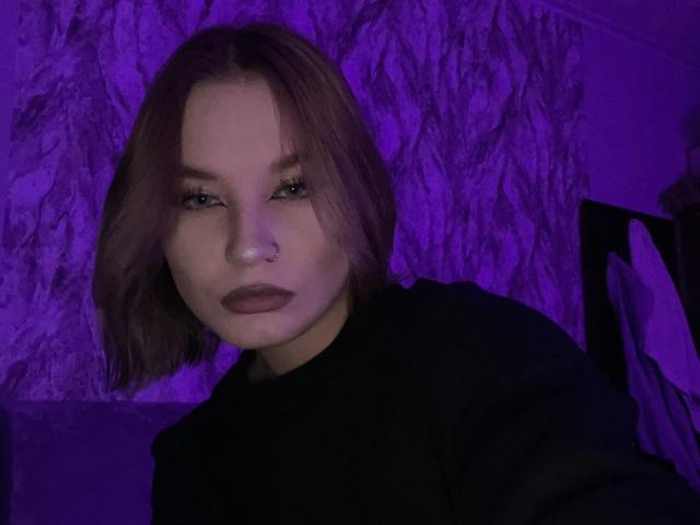 BertaVoltz mit einem violett schimmernden Gesicht, einem Nasenring und dunklem Lippenstift, trägt ein schwarzes Oberteil, hat kurze, braune Haare und blickt leicht zur Seite, in schwachem, violettem Licht..