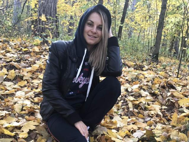 BelleLisaG, crouching with yellow leaves, wearing a black hoodie and jacket, black pants, smiling, blonde hair, hand in hood, autumn forest, soft light, relaxed, cute.