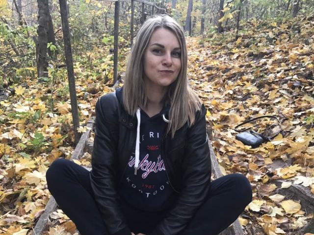 BelleLisaG, blonde, sitting cross-legged on wooden planks, wearing black leather jacket, black hoodie, black pants, autumn leaves covering ground, slight smile, casual, natural light, forest background.