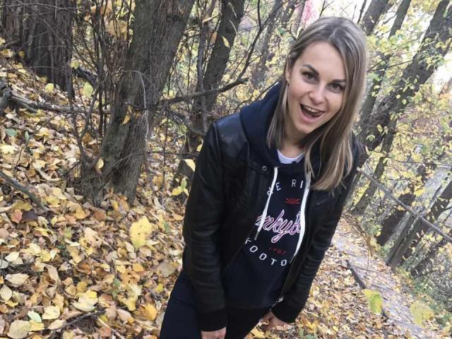 BelleLisaG with blonde hair, black leather jacket, and white hoodie, smiling, standing in forest with yellow leaves, hands by her sides, showing her tits slightly.