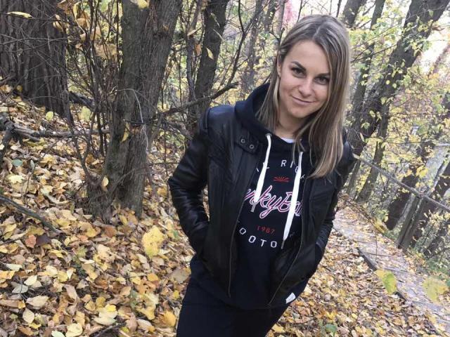 BelleLisaG with blonde hair, black leather jacket, and black hoodie standing in a forest with yellow leaves, hands in pockets, smiling slightly.