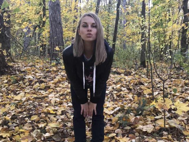 BelleLisaG with blonde hair, pouting lips, black hoodie, and jeans, bending slightly forward in a forest with yellow autumn leaves covering the ground.