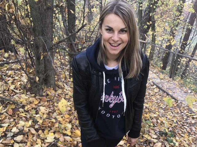 BelleLisaG with blonde hair, smiling, wearing a black leather jacket and "Adidas" hoodie, standing in a forest with yellow autumn leaves on the ground.