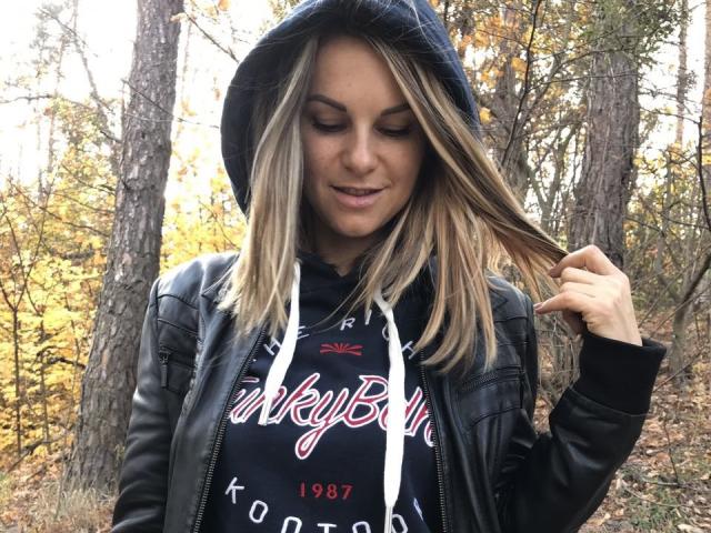 BelleLisaG with blonde hair, black hoodie, and leather jacket, holding hair, smiling, wearing a blue hat, autumn forest background, wearing a black "TinderBox" hoodie.