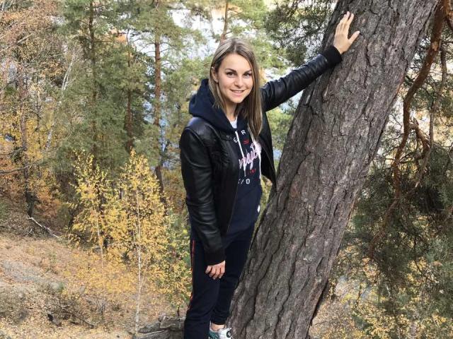 BelleLisaG with straight brown hair, black leather jacket, navy hoodie, and ripped jeans, standing against a tree in a forest, smiling, hand on trunk.