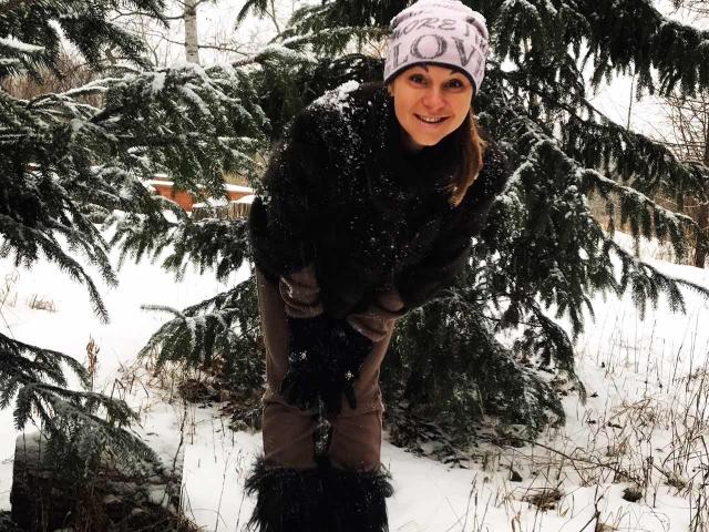 BelleLisaG bending forward in a snowy forest, wearing a white beanie, black fur coat, brown pants, black gloves, and boots, with a snow-covered evergreen tree in the background.