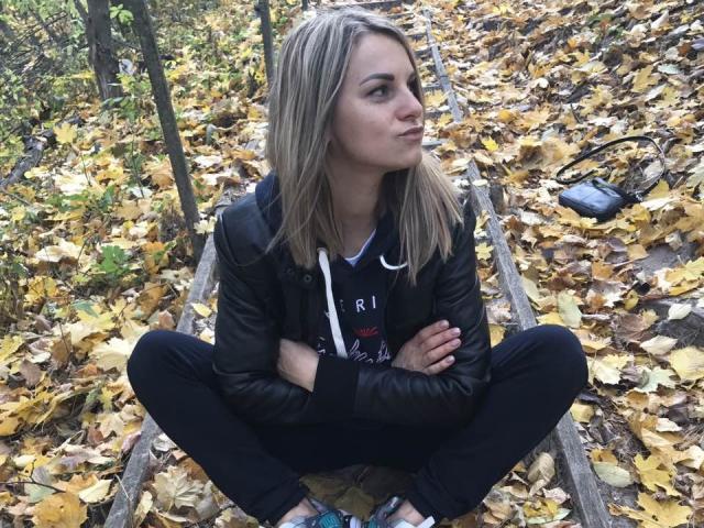 BelleLisaG, sitting on old wooden tracks with arms crossed, legs spread, wearing a black leather jacket and black pants, surrounded by yellow and brown autumn leaves.