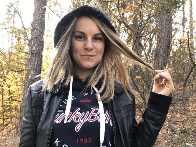 BelleLisaG with blonde hair, black leather jacket, and "bitchin" hoodie, holding her hair, smiling, in a forest with yellow leaves. She's got a cute face and small tits.