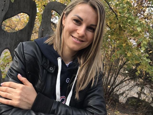 BelleLisaG with blonde hair, black leather jacket, and dark hoodie, smiling, arms crossed, standing in front of wooden fence with yellow-green leaves in background.