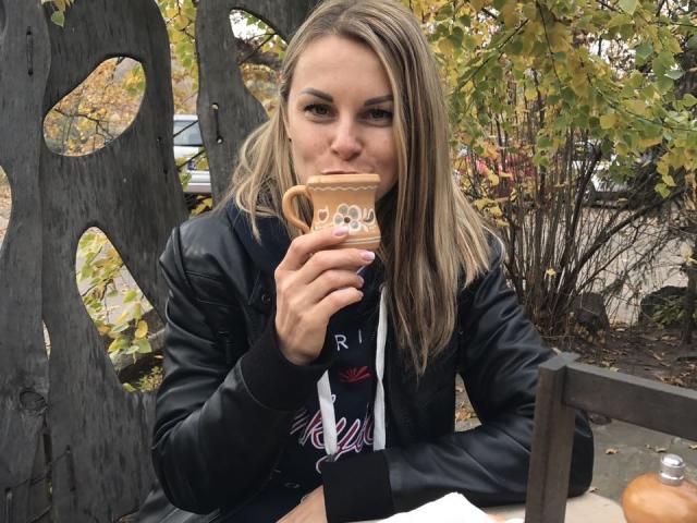 BelleLisaG with blonde hair, black leather jacket, sipping from a brown mug, sitting outside, autumn leaves in background, looking directly at camera with a slight smile.
