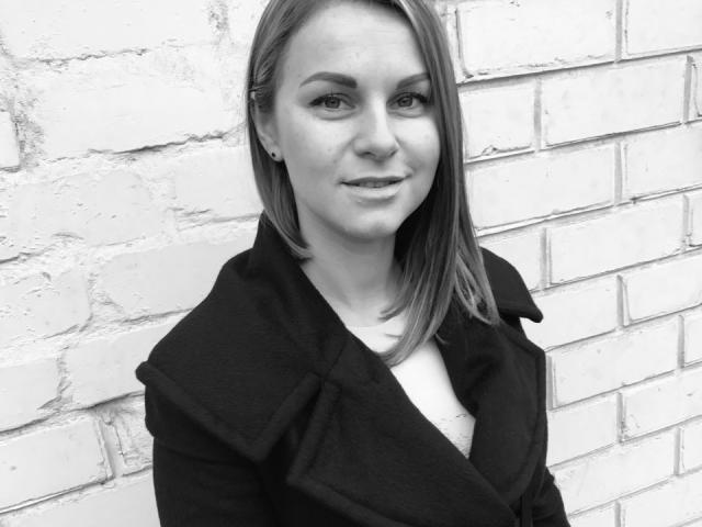 BelleLisaG has straight, medium-length hair, wearing a black coat, standing against a white brick wall, smiling slightly, with subtle makeup and small earrings. Black and white photo.