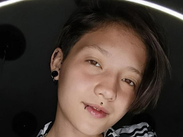 A LissyLuxe tem cabelo preto curto, olhos castanhos e um pequeno piercing no nariz. Ela está usando protetores auriculares pretos e uma camisa listrada, com um leve sorriso e um piercing no lábio..