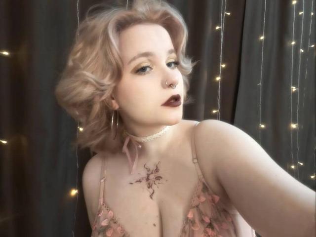 Mahalia Ryherd, con i capelli biondi e ricci, un anello al naso, rossetto scuro, un reggiseno floreale, una collana e un tatuaggio sul petto, guarda verso l'alto, con delle luci decorative sullo sfondo. Il seno è visibile, con una luce soffusa..