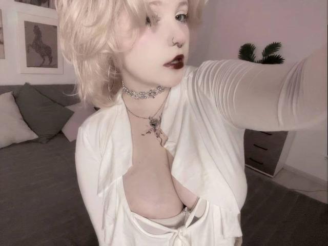 Mahalia Ryherd mit blonden Haaren, weißem Oberteil, tiefem Ausschnitt, der ihre Brüste zeigt, rotem Lippenstift, Nasenring, Choker und einer tätowierten Halskette, macht ein Selfie in einem schwach beleuchteten Raum..