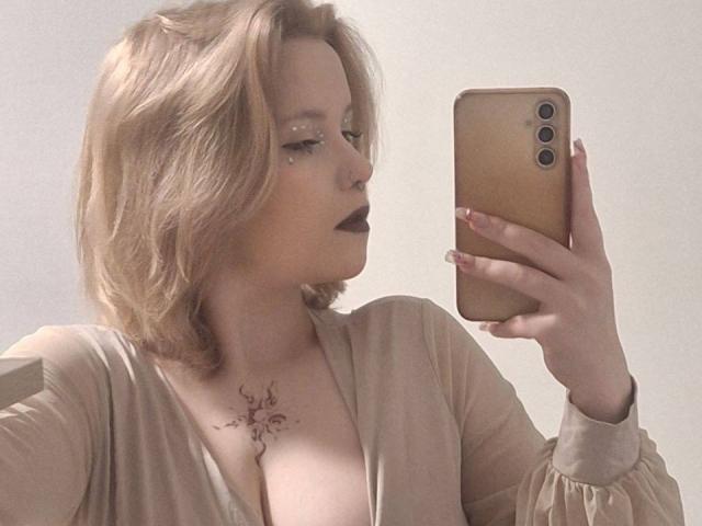 MahaliaRyherd, met blond haar en donkere lipstick, maakt een selfie in een spiegel. Ze heeft een tatoeage op haar borst en draagt een beige top, terwijl ze een gouden telefoon vasthoudt..