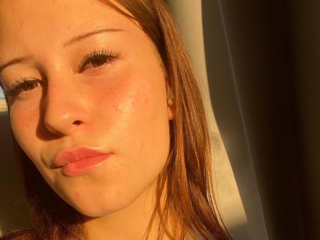 SharenMcdale's face, sunlit, with light brown skin, subtle freckles, and pink pouty lips, has long brown hair, and thick, long eyelashes, looking slightly to the side.