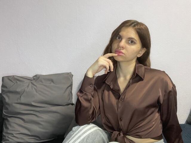 CicelyDence with brown shirt tied at waist, grey pants, and finger near pink lips, sitting on grey pillow. She has light skin, brown hair, and slightly confused expression.