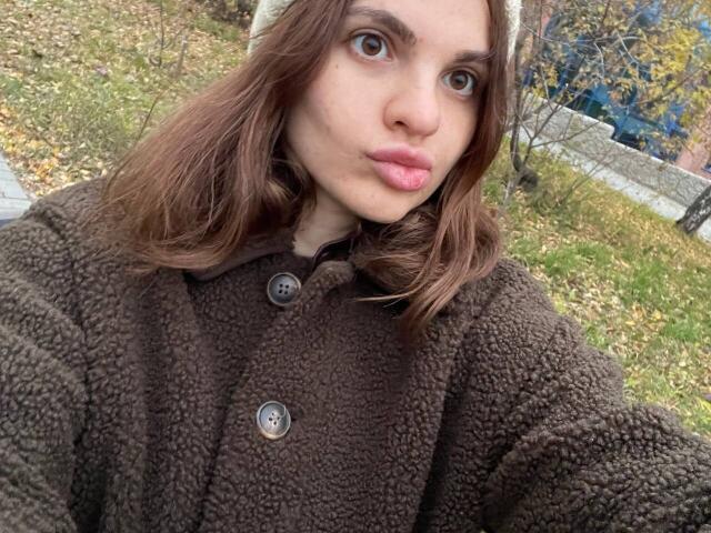 CicelyDence has pouty pink lips, brown wavy hair, and wears a fluffy brown coat with buttons, a white beanie, and looks directly into the camera with wide eyes.