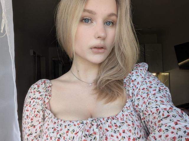 RealAnna with blonde hair, blue eyes, and fair skin, wearing a floral dress, showing her tits in a soft light, looking straight at the camera with a neutral expression.