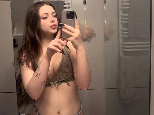 MaidaCain taking a mirror selfie, wearing a brown lace bralette, showing her tits, with tattoos on her left arm and a pouty face, holding a phone with a black case.