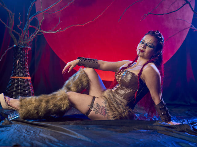 Cataleya, une jeune femme aux cheveux roux tressés, portant un corset en cuir, des jambières en fourrure et des tatouages, assise sur un tissu noir, les bras en arrière et les jambes croisées, éclairée par des lumières rouges et bleues, les bras ornés de dentelle noire..