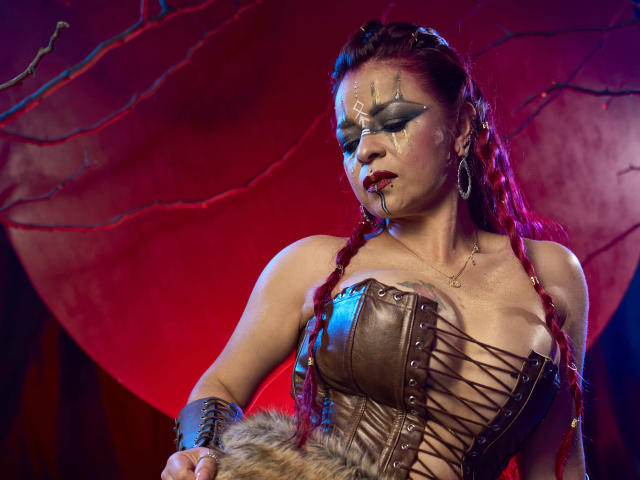 Cataleya Sweett, avec des tresses rousses, du maquillage de guerre gris, un corset en cuir laissant entrevoir de grandes seins, tenant une fourrure, éclairage rouge et bleu, regardant sérieuse, portant des boucles d'oreilles pendantes, provenant de Brazzers..