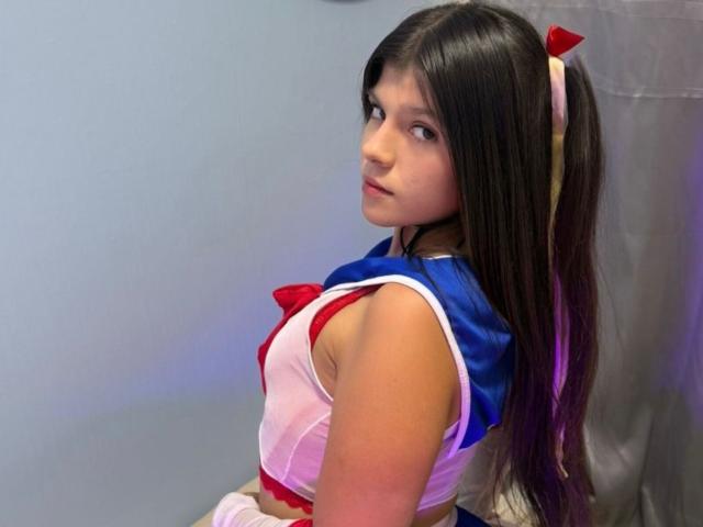 RilleyFoyx with long brown hair, blue and white cosplay outfit, red ribbon, looking back, showing small tits, slim, cute, wearing a pink hairband, in a simple indoor setting.