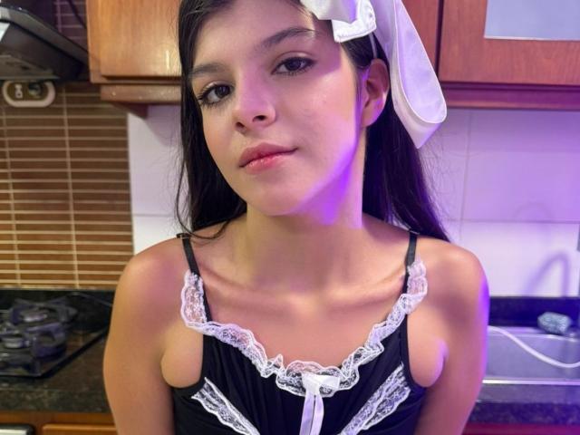 RilleyFoyx with long black hair, white bow, black lace-trimmed top, showing small bare tits, standing in kitchen, purple light on right side.