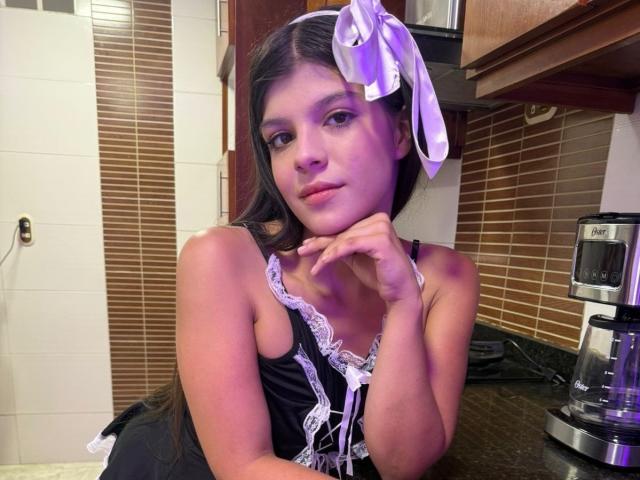 RilleyFoyx with long black hair, purple ribbon, black lace-up top, resting chin on hand, looking at camera, kitchen background, coffee maker on right, brown tile wall.
