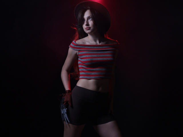 JasmineNeal with dark hair, wearing a red-striped off-shoulder top, black shorts, and a fingerless glove, stands against a dark background with red lighting highlighting her face.